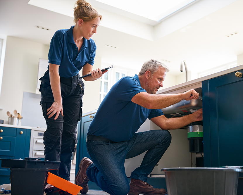 a plumber works under a sink while another plumber supervises